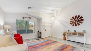 Ein geräumiges Wohnzimmer mit großem Fenster, einem farbenfrohen Teppich, einem weißen Konsolentisch mit Pflanzen und einem dekorativen Wandbild.