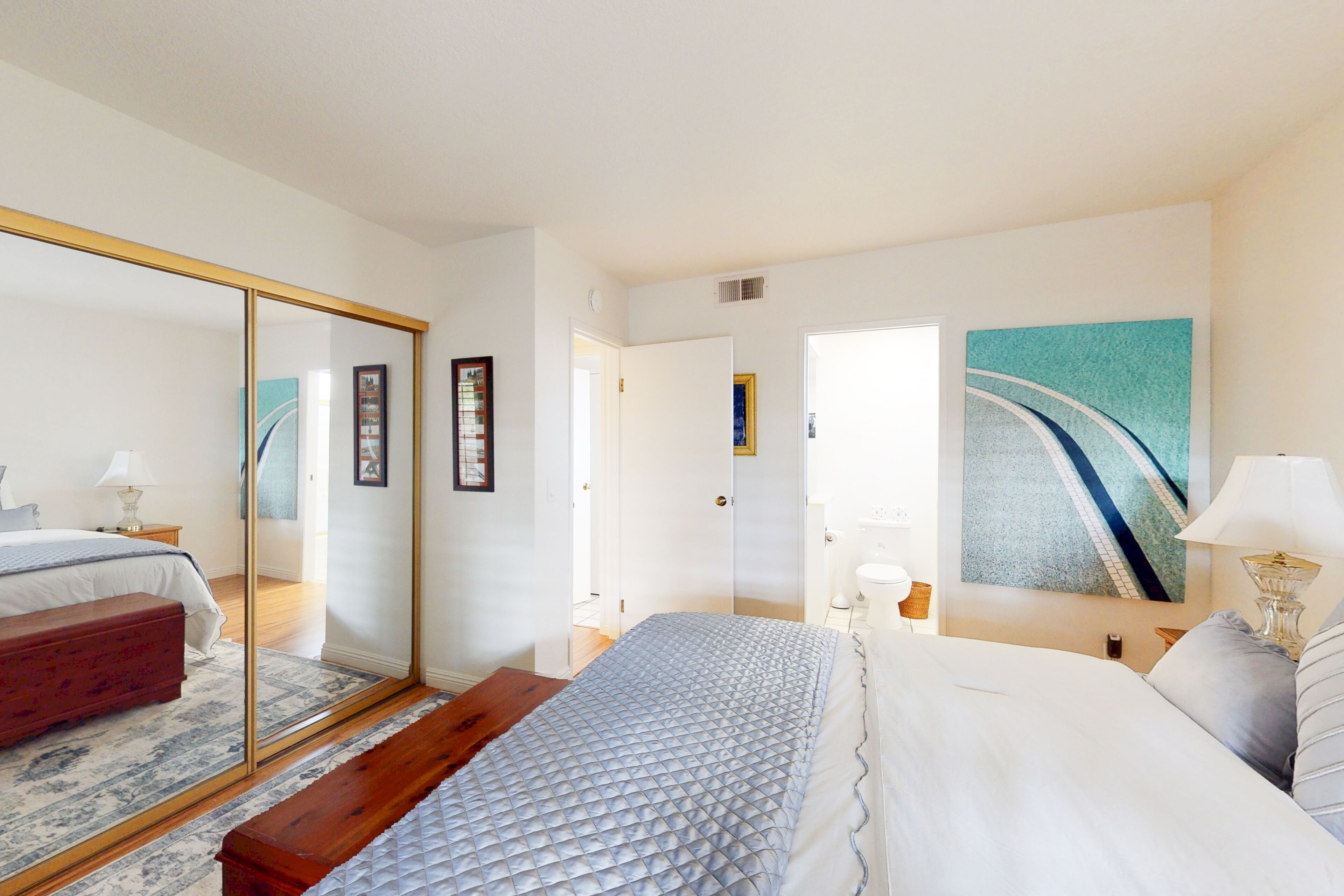 A bedroom with a bed, a mirror, a painting, and a wooden bench.