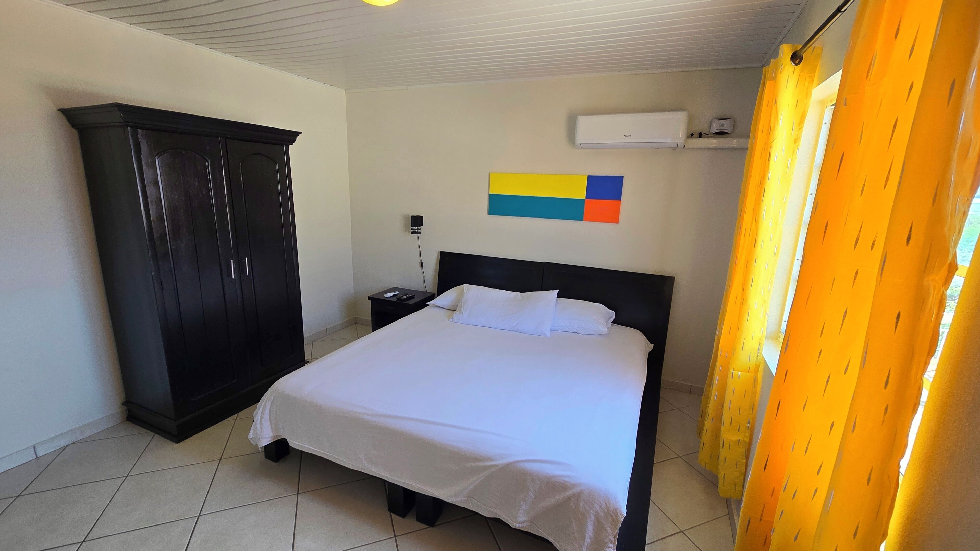 A hotel room with a bed, a wardrobe, a nightstand, an air conditioning unit, and a colorful painting on the wall.