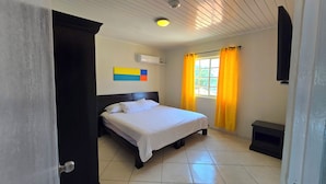 A hotel room with a bed, a window with yellow curtains, a ceiling fan, and a wall-mounted air conditioner. - Adorable 1-bedroom Apartment 3 km From Eagle Beach (Noord)
