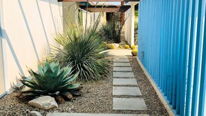 A narrow pathway with stepping stones leading to a covered area, surrounded by various desert plants and a blue fence. - Cheerful mid-century home w/ private pool, spa, firepit & mountain views (Palm Springs)