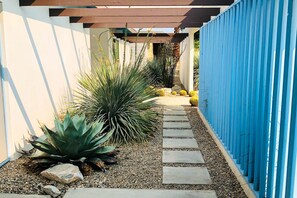 A narrow pathway with stepping stones leading to a covered area, surrounded by various desert plants and a blue fence. - Cheerful mid-century home w/ private pool, spa, firepit & mountain views (Palm Springs)