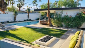 A backyard with a swimming pool, a covered seating area, and a well-maintained lawn. - Cheerful mid-century home w/ private pool, spa, firepit & mountain views (Palm Springs)