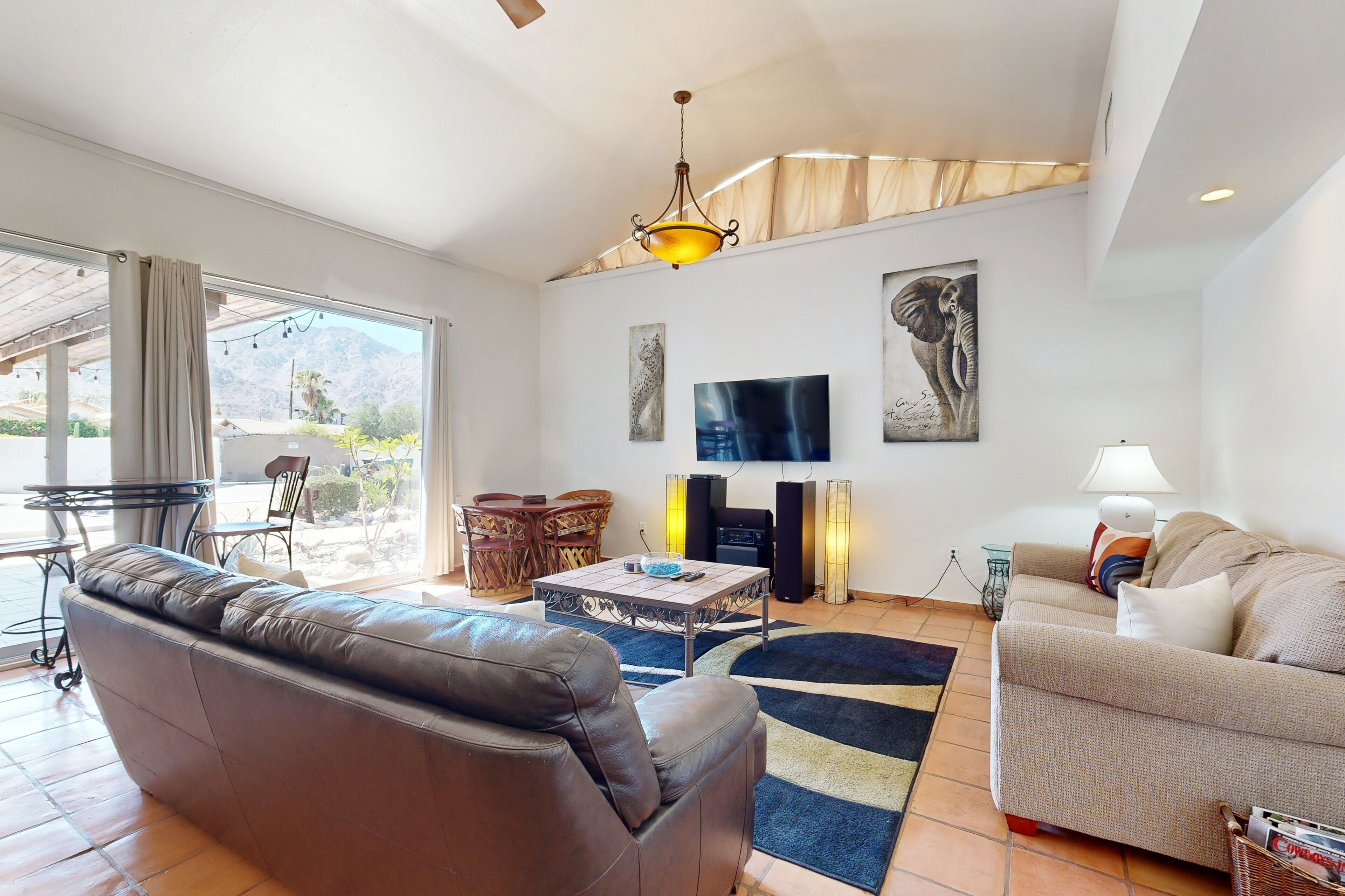 Un salon avec un canapé en cuir marron, un canapé beige, une table basse et un téléviseur. Il y a deux tableaux au mur, une lampe et une fenêtre avec des rideaux.