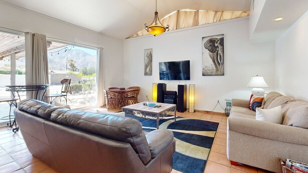 Ein Wohnzimmer mit braunem Ledersofa, einem beigefarbenen Sofa, einem Couchtisch und einem Fernseher. An den Wänden hängen zwei Gemälde, eine Lampe steht dort und ein Fenster ist mit Vorhängen drapiert.