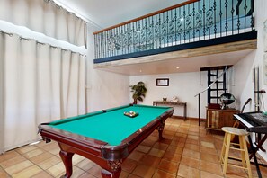 Une table de billard avec des billes, un piano et un tabouret dans une pièce dont le sol est carrelé, et un balcon agrémenté d’une balustrade décorative.