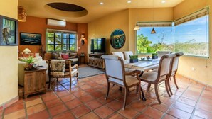 A spacious living area with a dining table, chairs, and a TV. There are large windows offering a view of the outdoors. - 3 Bedroom with Pool & Mountain Views (Los Barriles)