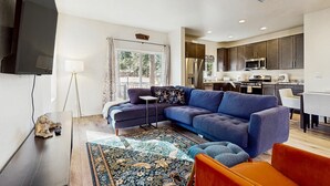 A modern living room with a sectional sofa, a flat-screen TV, and a kitchen in the background. - Modern, dog-friendly bungalow home with patio, balcony, & central AC (Flagstaff)