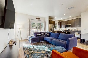 A modern living room with a sectional sofa, a flat-screen TV, and a kitchen in the background. - Modern, dog-friendly bungalow home with patio, balcony, & central AC (Flagstaff)