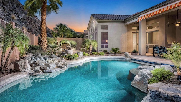 A swimming pool with a curved edge, surrounded by rocks and palm trees, with a building in the background.