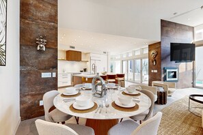 Un espace repas moderne avec une table ronde dressée pour quatre personnes, une cuisine en arrière-plan et un grand téléviseur fixé au mur.