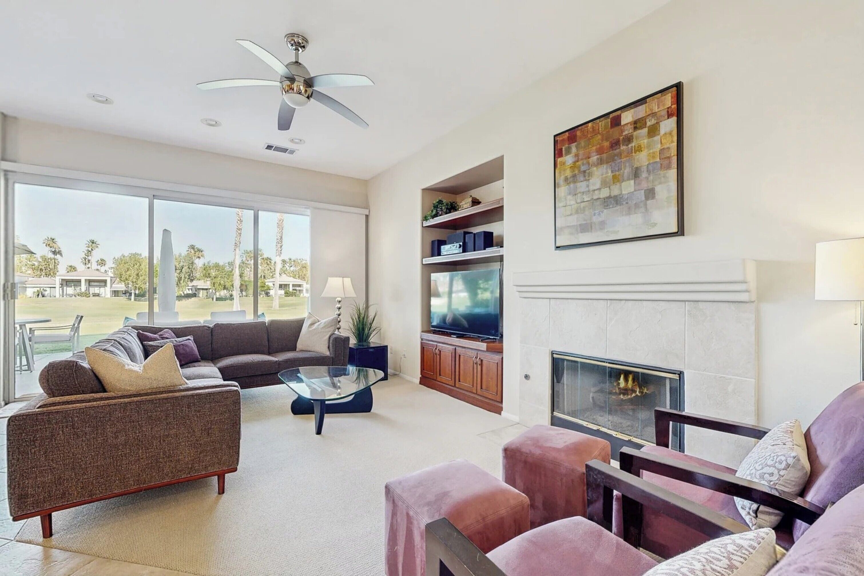 A spacious living room with a large window, a ceiling fan, a fireplace, and a television.