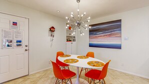 Ein Essbereich mit einem weißen Tisch, orangefarbenen Stühlen, einem modernen Kronleuchter, einem Gemälde an der Wand und einer Tür mit Infografiken.