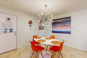 A dining area with a white table, orange chairs, a modern chandelier, a painting on the wall, and a door with informational posters. - Ground-floor condo with private hot tub, 8 on-site pools, & tennis/pickleball (Palm Springs)