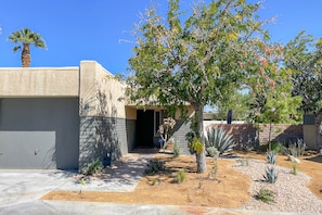 A modern building with a covered entrance, a small garden with various plants, and a palm tree. - Ground-floor condo with private hot tub, 8 on-site pools, & tennis/pickleball (Palm Springs)