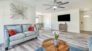 A living room with a grey sofa, red and orange pillows, a wooden coffee table with a vase of white flowers, a ceiling fan, and a flat-screen TV mounted on the wall. - Single-level dog-friendly home with private pool, grill, enclosed backyard, & AC (Chandler)