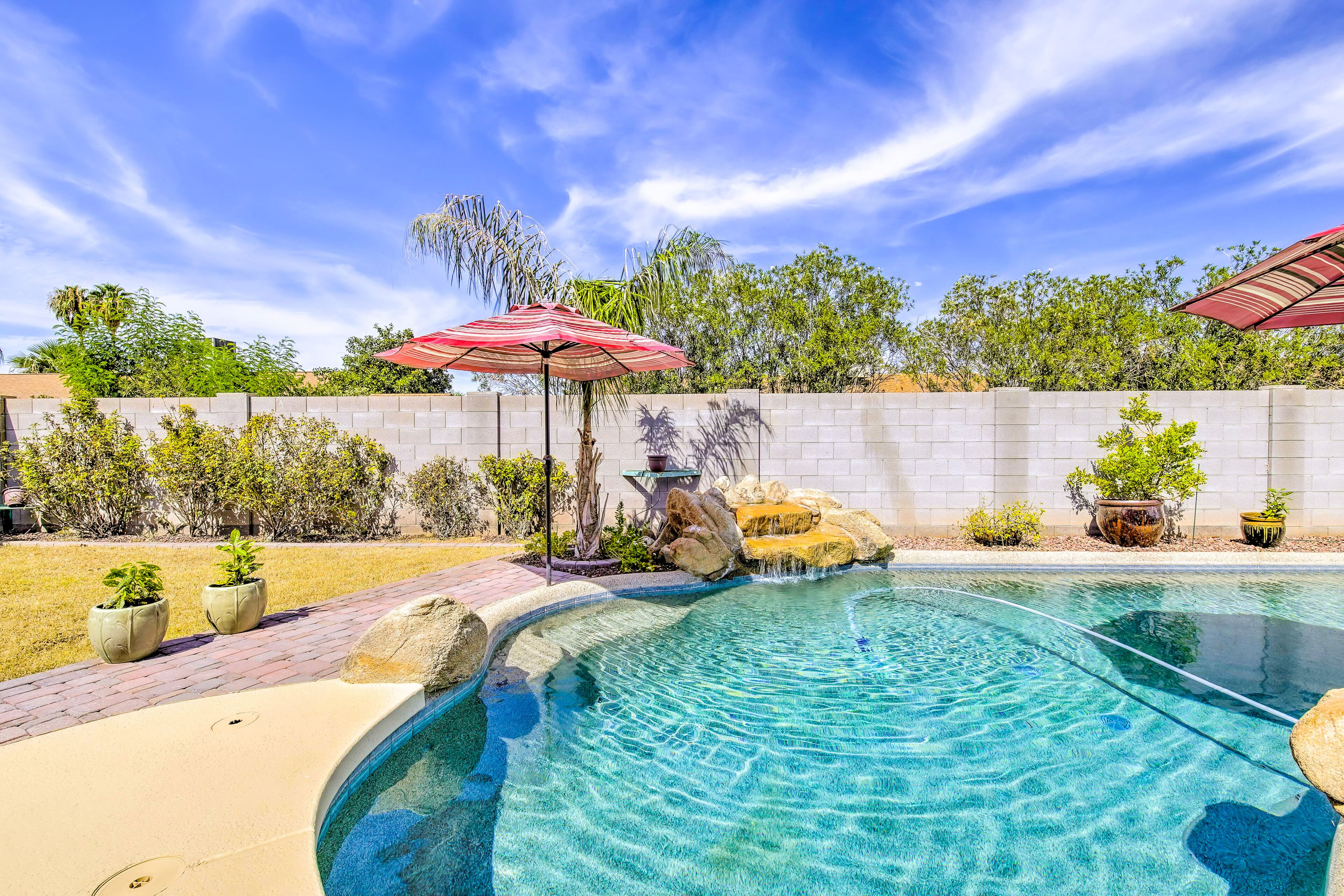 A clear swimming pool with a stone border, surrounded by a brick patio, a white wall, and various potted plants.
