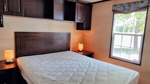 Ein Schlafzimmer mit einem Bett, einem Nachttisch mit Lampe, einem Fenster mit Blick auf Bäume und dunklen Holzschränken über dem Bett.