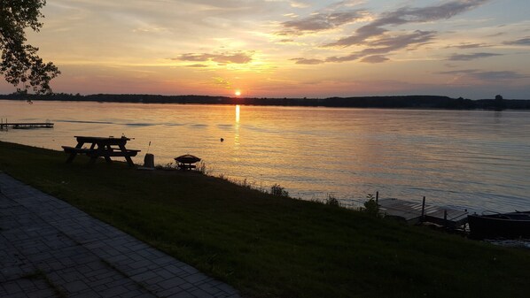 A lakeside view with a picnic table and grill, calm water, and a sunset.
