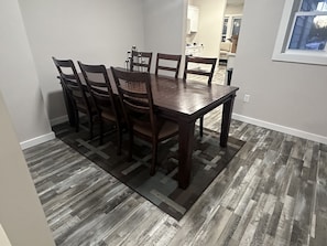 A dining area with a wooden table and chairs, a patterned rug, and a kitchen in the background. - State Street Retreat (Big Rapids)