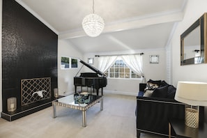 A living room with a grand piano, a glass coffee table, a black sofa, a chandelier, and a fireplace. - Sunshine, Style & Your Own Private Pool Retreat – Dog-Friendly!- #112181 (La Quinta)