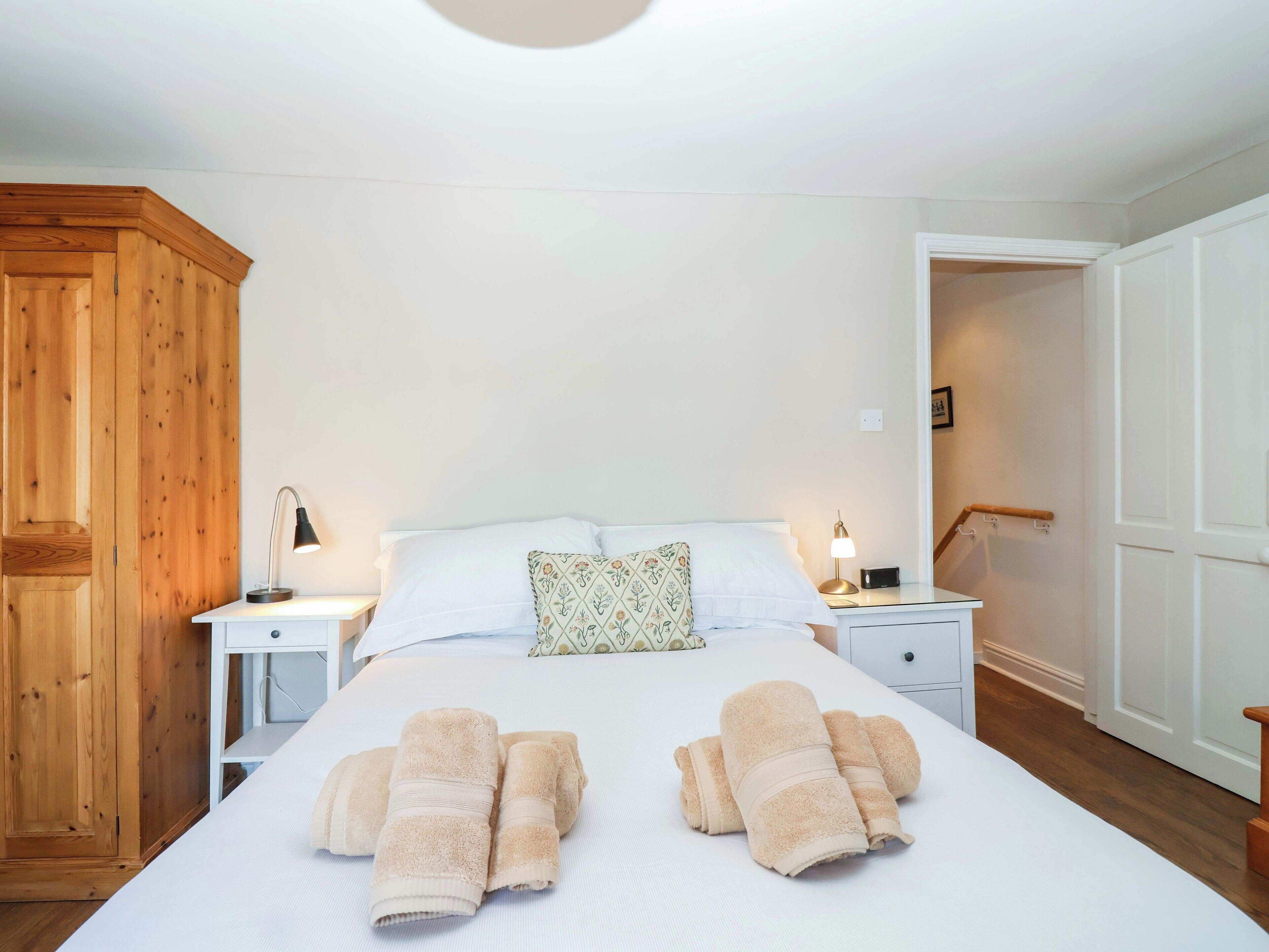 A neatly made bed with rolled towels, two bedside tables with lamps, a wooden wardrobe, and a white door.