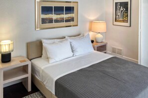 A neatly made bed with white bedding and a gray blanket, a bedside lamp, and a framed picture on the wall.