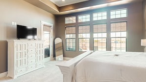 Een slaapkamer met een groot bed, een televisie op een witte dressoir en meerdere ramen.