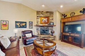 A living room with a stone fireplace, leather furniture, a large flat-screen TV, and world maps on the wall. - Luxurious home with an outdoor kitchen, private pool & spa - close to golf (Bermuda Dunes)