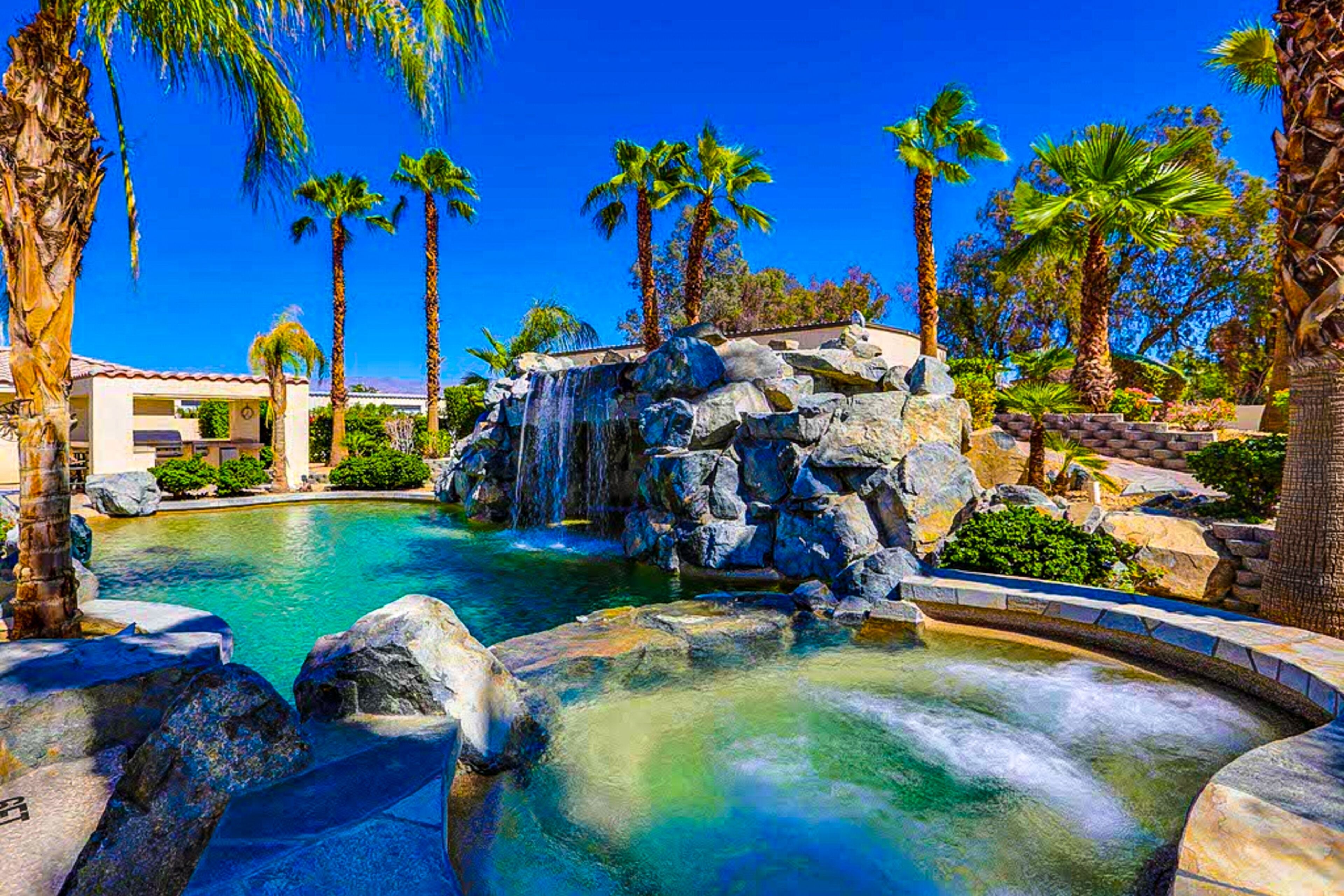 A pool with a waterfall, surrounded by palm trees and a building with a red roof.