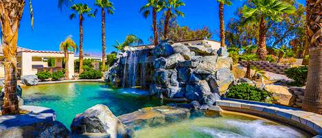 A pool with a waterfall, surrounded by palm trees and a building with a red roof.