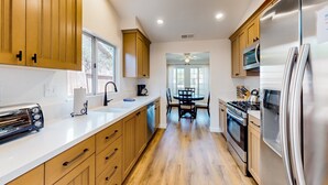 A modern kitchen with wooden cabinets, stainless steel appliances, a toaster, and a dining area with a table and chairs. - Dog-Friendly Home w/ a Private Pool, Pool Spa, Firepit, & Patio - 3BR - #66716 (La Quinta)