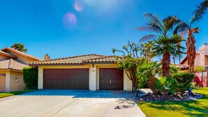 A residential house with a tiled roof, two garage doors, and a well-maintained lawn with palm trees. - Dog-Friendly Home w/ a Private Pool, Pool Spa, Firepit, & Patio - 3BR - #66716 (La Quinta)