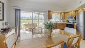 A modern kitchen with wooden cabinets, a stainless steel refrigerator, and a dining area with a glass door leading to a patio. - Sun, Comfort, and Stunning Views, Shared pool & Dog-friendly (Palm Desert)