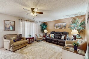 A living room with a beige sofa, a brown leather couch, a beige armchair, and a coffee table. There is a ceiling fan, a large plant, and two paintings on the walls. - Sunny home with covered patio, grill, pool, tennis, hot tub, full kitchen, & AC (Mesa)