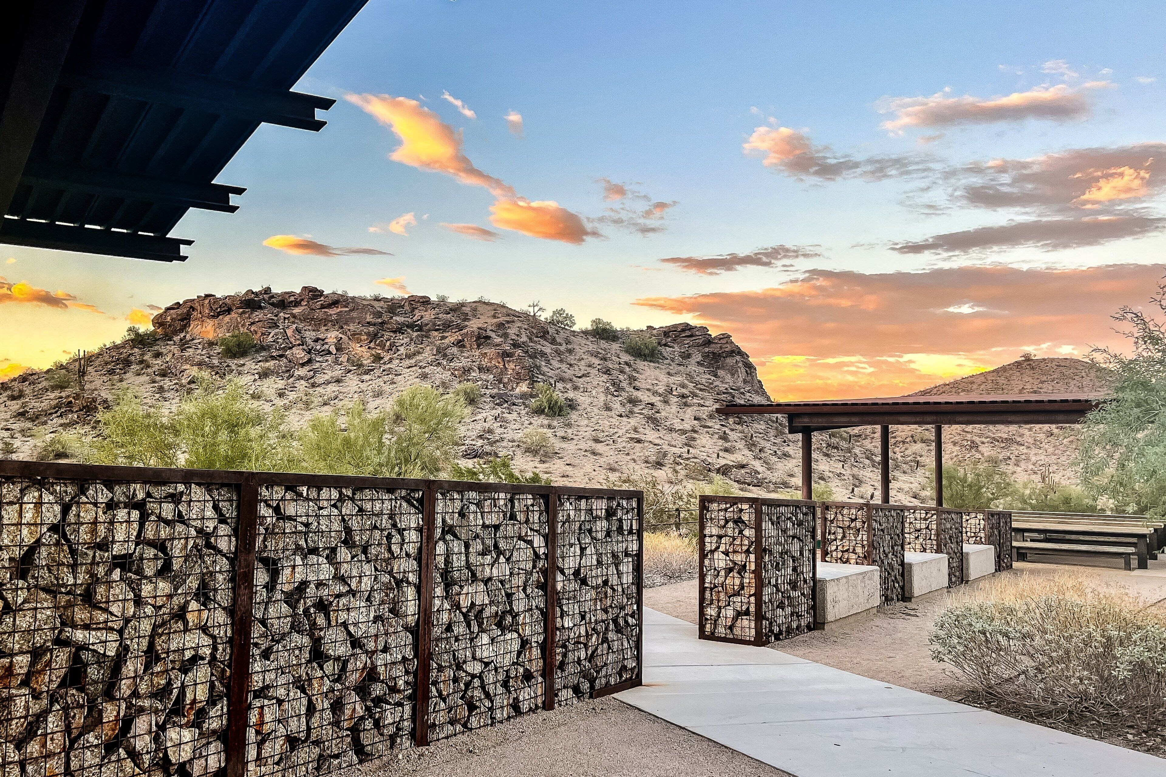 A desert landscape with a rocky hill, a wooden pavilion, and a stone fence.