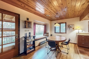 Una habitación acogedora con techo de madera, una mesa de comedor con sillas, una estantería y una ventana con cortinas.