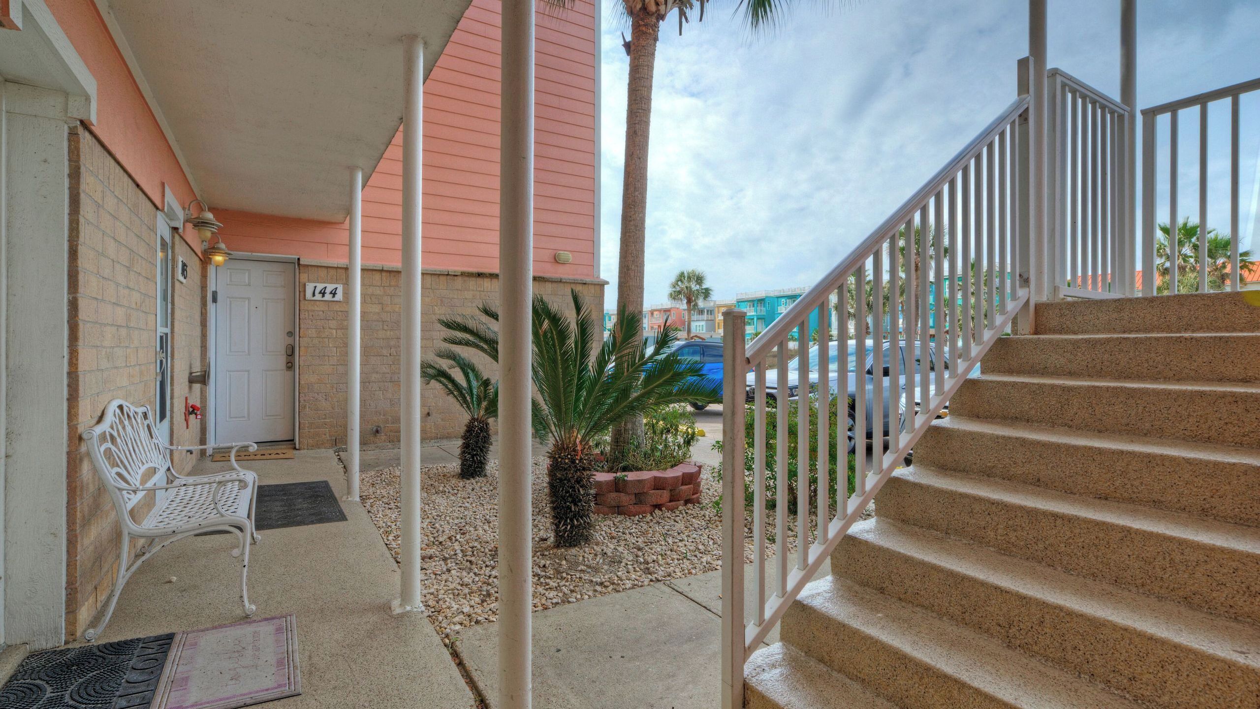 A building with a number '144' on the door, a covered porch with a bench, a palm tree, and a staircase.