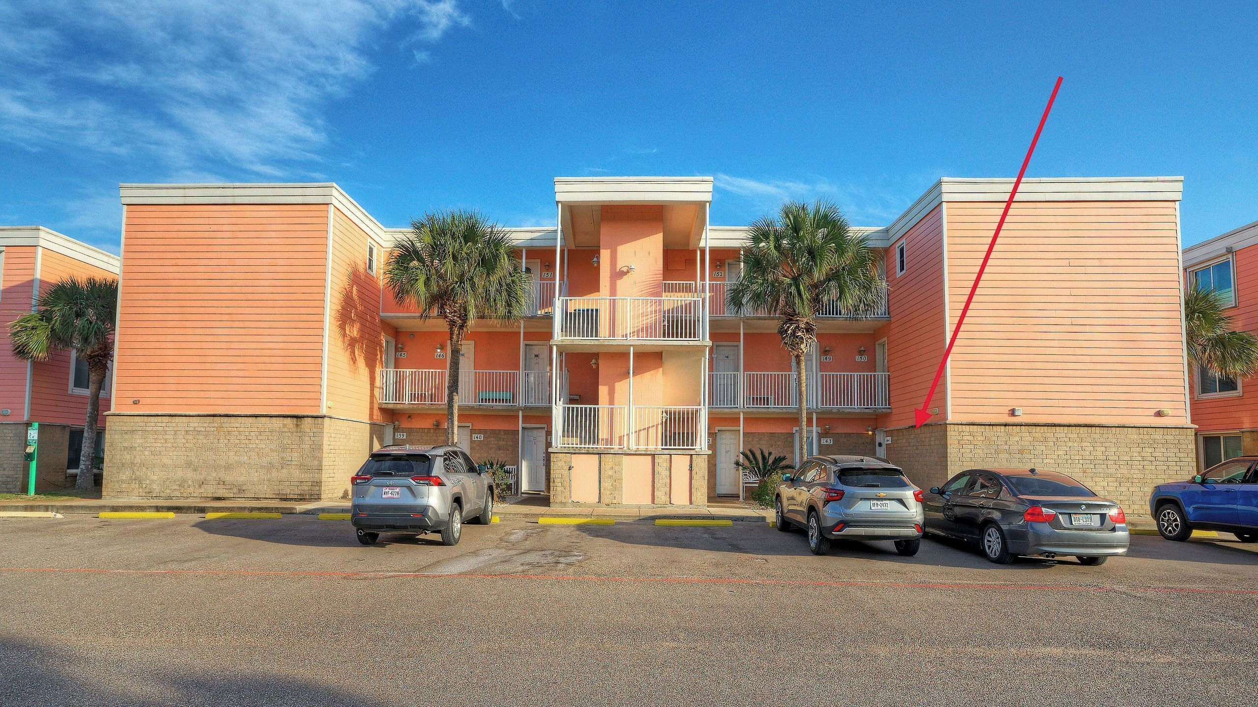 A multi-story building with balconies, parking in front, and palm trees.