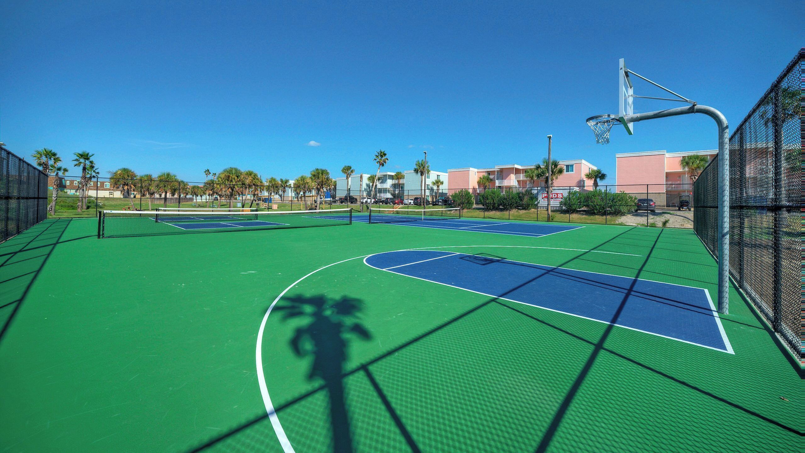 A multi-sport court with a basketball hoop, surrounded by a chain-link fence, under a clear blue sky.