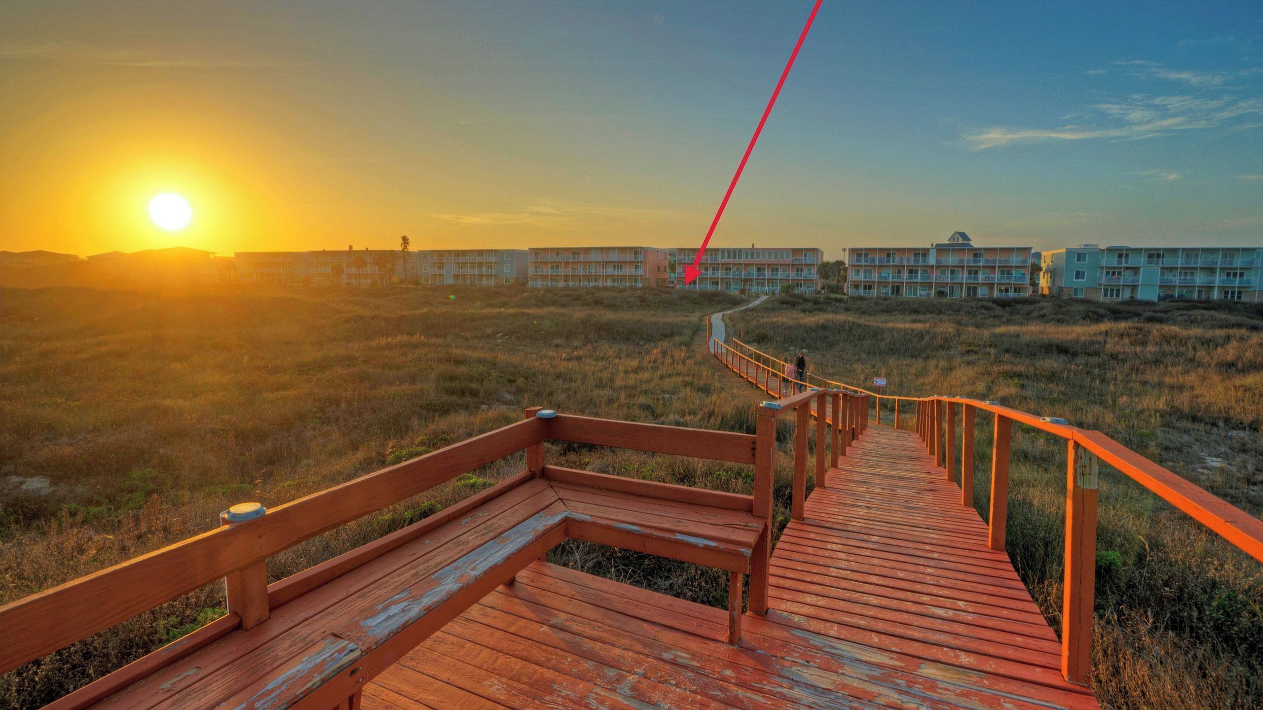 En træpromenade, der fører hen til et strandhotel med flere bygninger i baggrunden.
