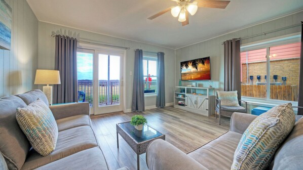 A living room with a sofa set, a coffee table with a plant, a TV, and a balcony with curtains. - Rest Ashored by Avantstay Port A Condo + Beach Access + Pickleball Court + Basketball Court (Port Aransas)