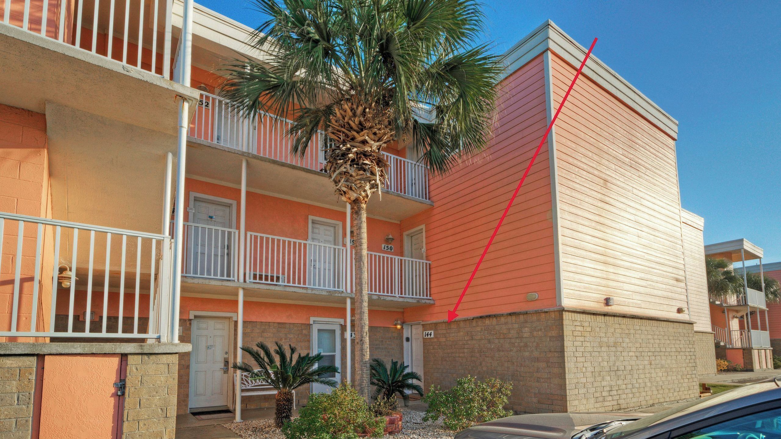 A multi-story building with balconies, a palm tree, and a car parked in front.
