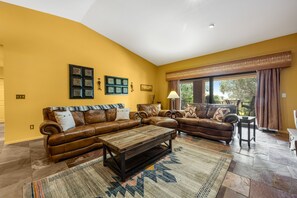 Ein Wohnzimmer mit einem braunen Ledersofa, einem Holztisch und einem gemusterten Teppich. Zwei Bilderrahmen sind an der Wand angebracht und eine Stehlampe steht im Raum.