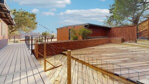 Una terraza de madera con un muro de ladrillo y un área cercada, rodeada de árboles y un cielo despejado.