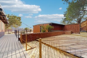 Eine Holzterrasse mit einer Backsteinmauer und einem eingezäunten Bereich, umgeben von Bäumen und blauem Himmel.