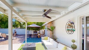 En täckt uteplats med en vit soffa, ett soffbord och en takfläkt. I bakgrunden finns en liten pool, och utemöbler syns.