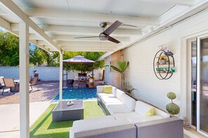 Ein überdachter Patio-Bereich mit einem weißen Sofa, einem Couchtisch und einem Deckenventilator. Im Hintergrund ist ein kleiner Pool zu sehen, und Gartenmöbel sind sichtbar.