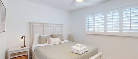 A bedroom with a bed, nightstand, bedside table, and a window with blinds.