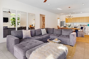Ein modernes Wohnzimmer mit einem grauen Ecksofa, einem Couchtisch und einer Felldecke. Es gibt einen Essbereich mit einem Holztisch und Stühlen, sowie eine Küche mit Holzschränken und Edelstahlgeräten.